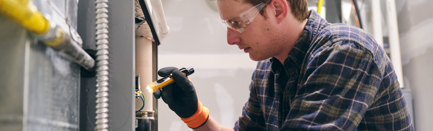 A man holding a flashlight to inspect a furnace.