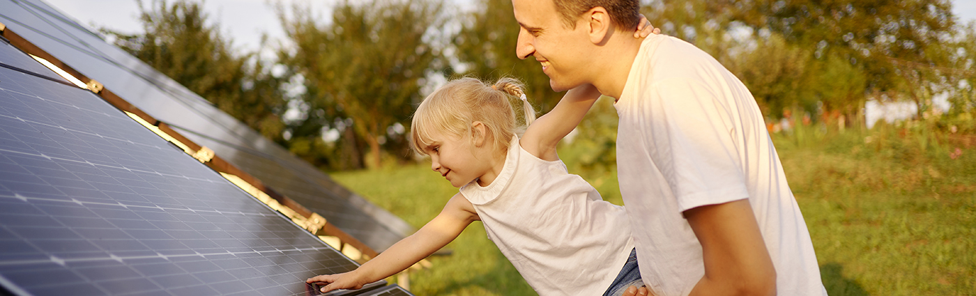 A man holding his daughter while she puts a hand on a solar panel.