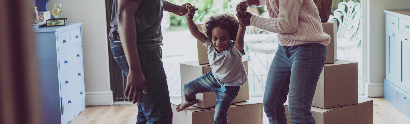 A father and mother each holding their child's hand and lifting her up in a living room, standing next to moving boxes.