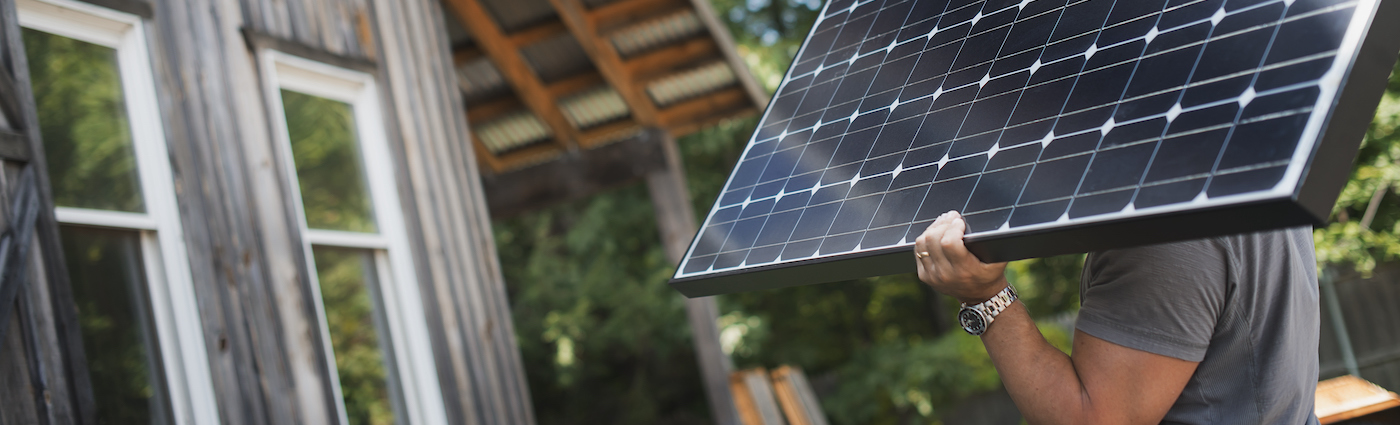 A man carrying a black solar panel and walking towards a home.