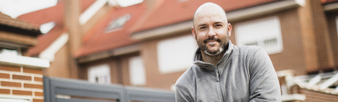 A smiling, bald businessman wearing a jacket standing in front of a brick house.