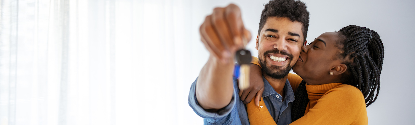 A woman in orange sweater kissing a man on the cheek while he's smiling and holding a key to his home.