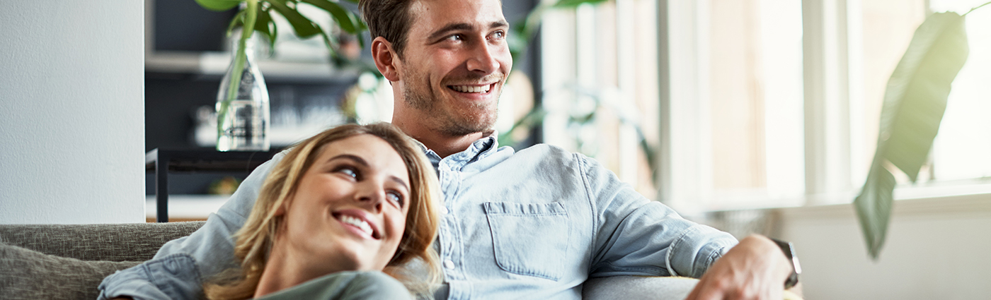 A man and woman sitting on a couch in a living room, smiling and looking out the window.