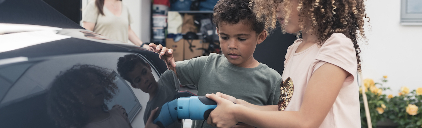 A girl plugging a charger into an electric vehicle while her sibling stand and watch.