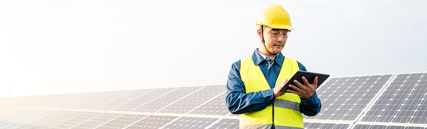 A worker wearing a yellow helmet and vest standing next to solar panels and looking at his iPad.