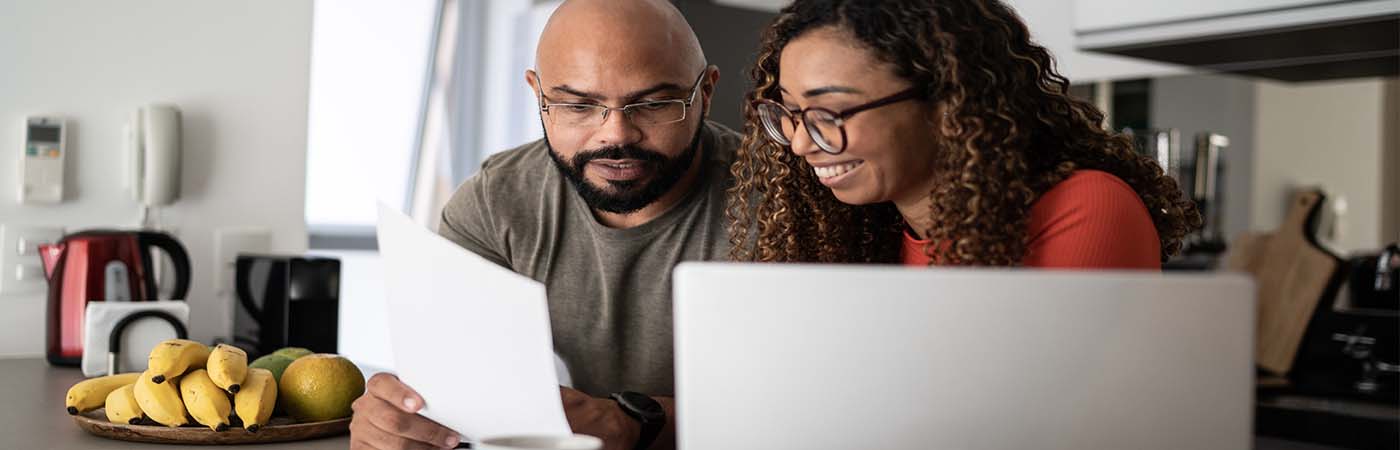 A couple sitting in front of a lap top while looking at and talking about a paper in the man's hand.
