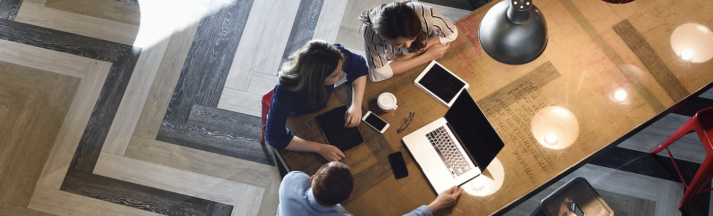 A group of people sitting around a wooden desk and looking at a laptop.