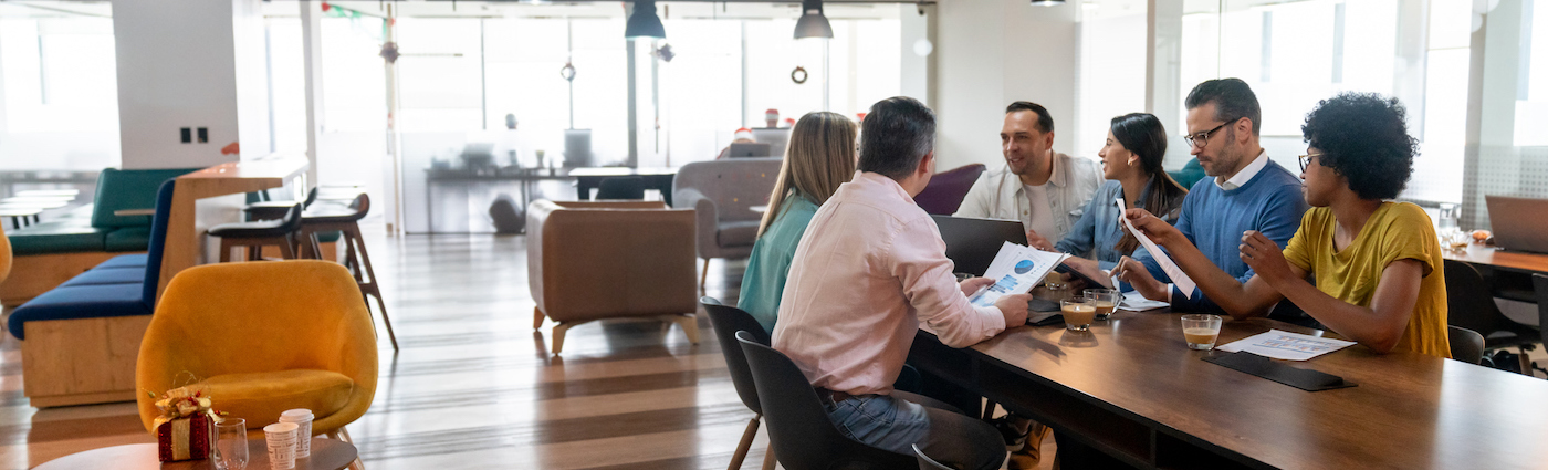 A group of six people sitting at a desk in an office talking and looking at some statistics on papers.