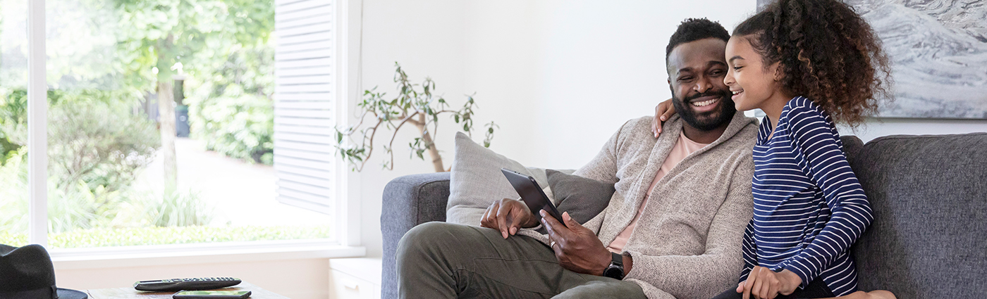 A father holding an iPad sitting with his daughter on a couch in a living room.