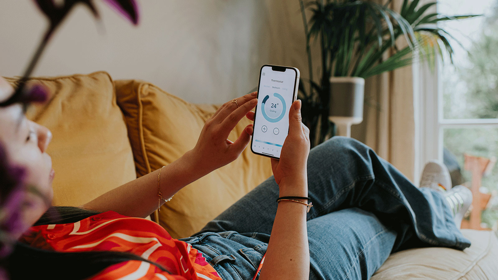 A homeowner lays on a yellow couch at home, checking their smart thermostat app on a smart phone.